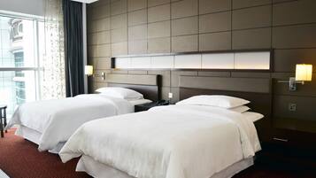 A hotel room with two beds, a large window with curtains, a modern headboard, and wall-mounted lamps.