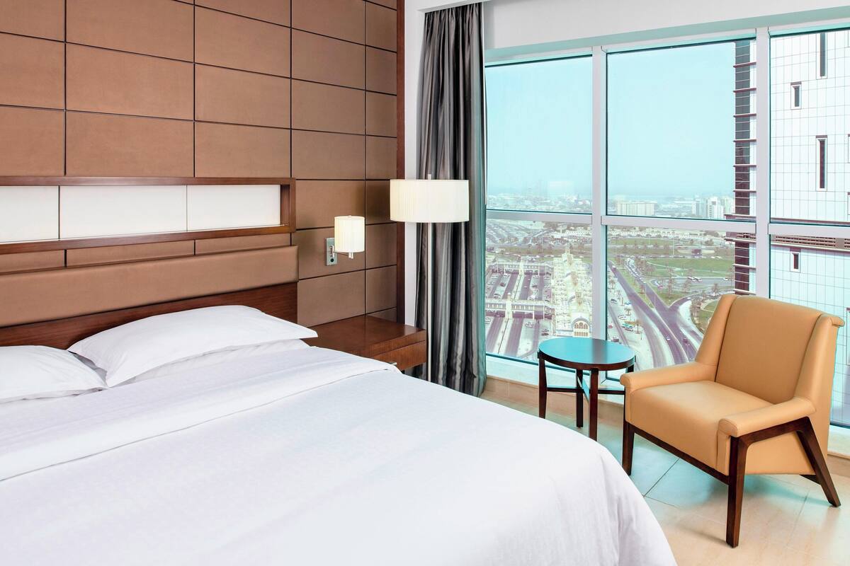 A hotel room with a large bed, a chair, a small table, and a view of a cityscape through large windows.