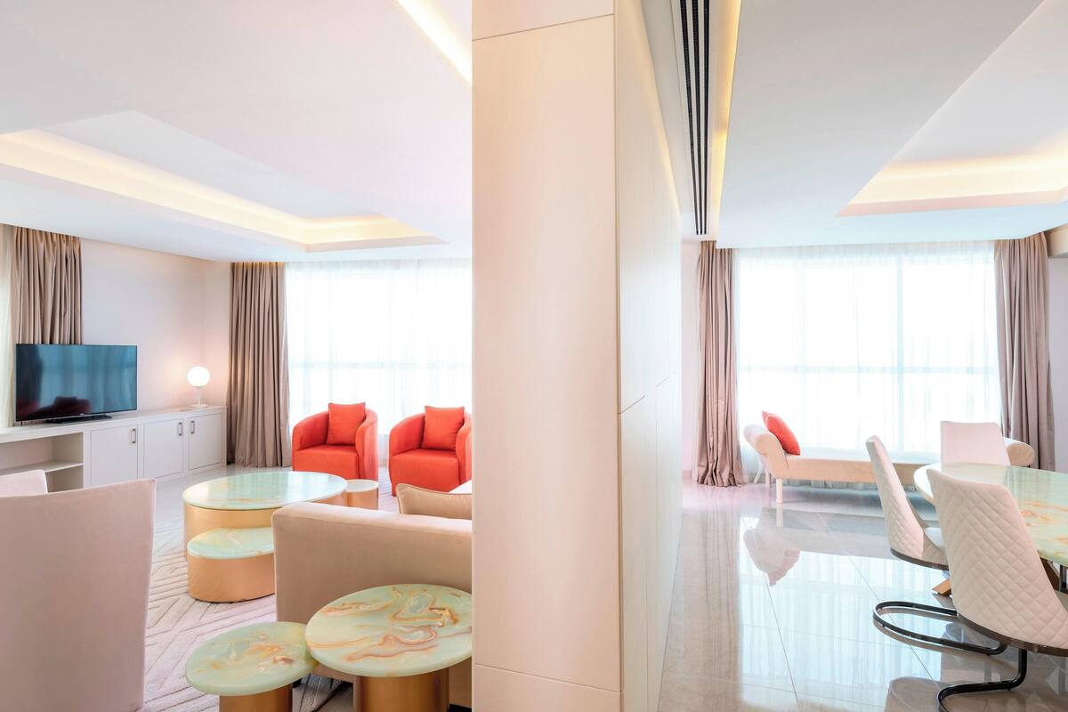 A modern hotel room with a flat-screen TV, a sofa, a dining table with chairs, and a large window with curtains.