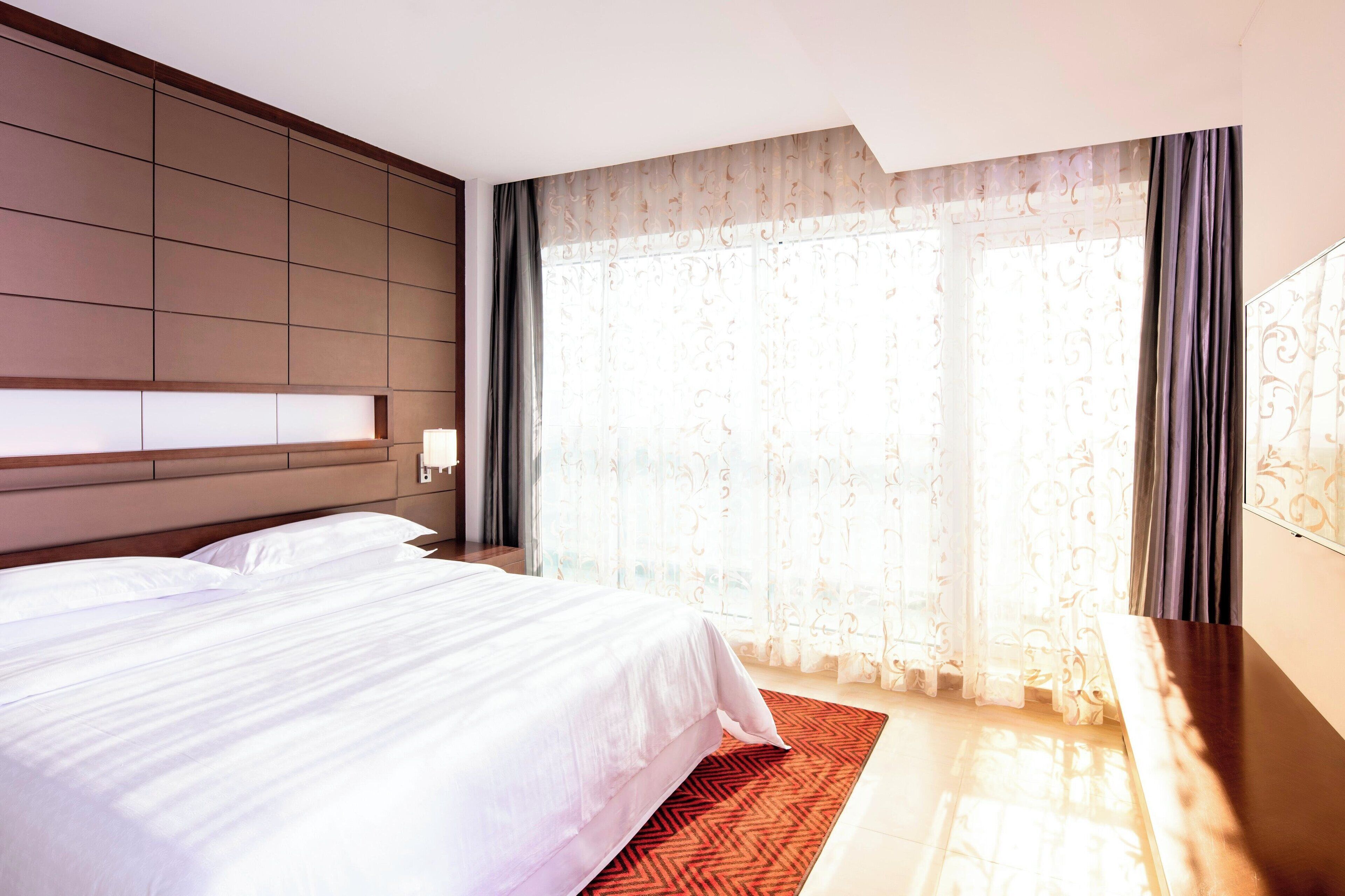 A hotel room with a large bed, a wooden headboard, a nightstand, and a large window with curtains.