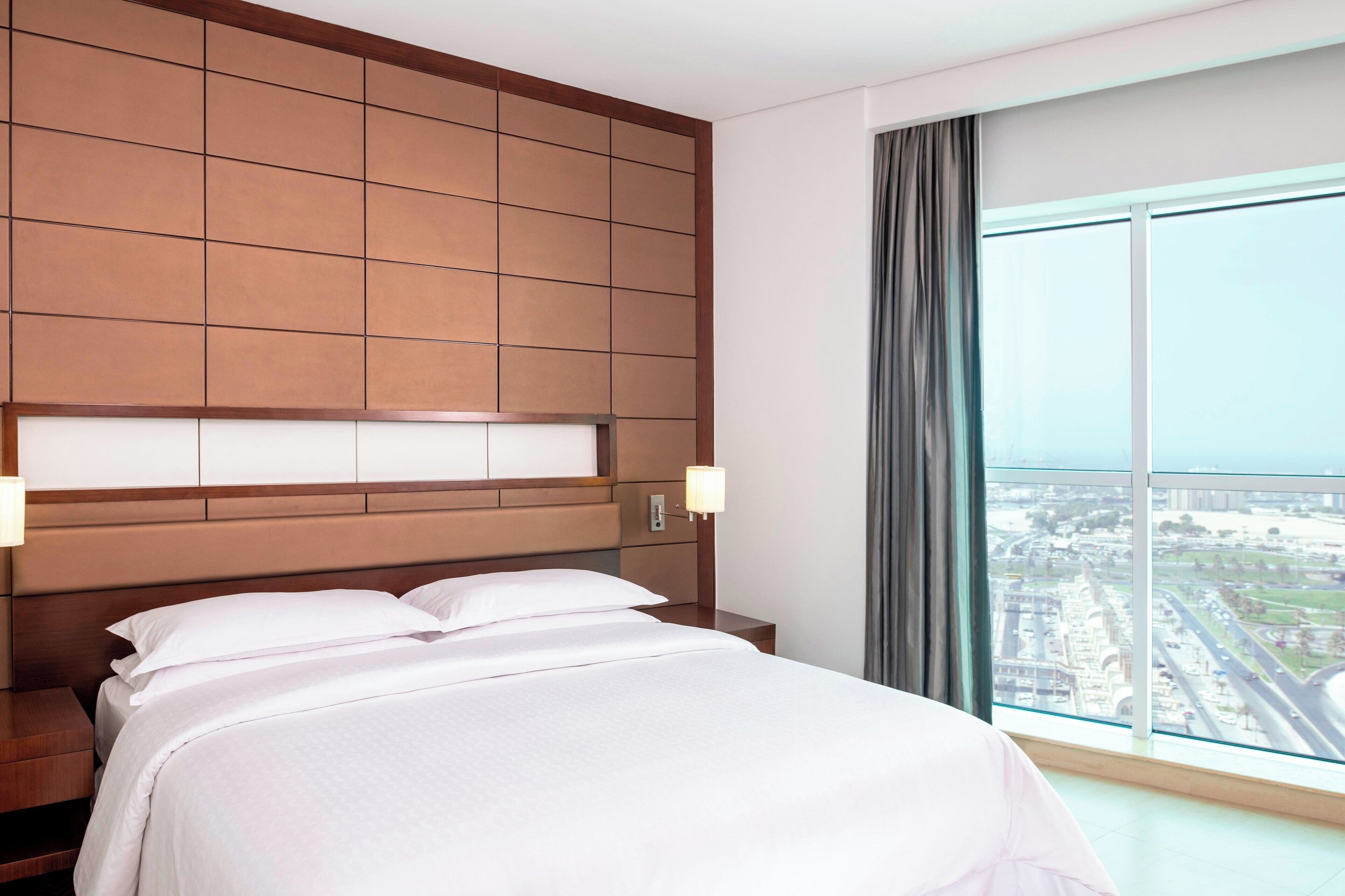 A hotel room with a large bed, a wooden headboard, a nightstand, a lamp, and a large window with a view of a cityscape.