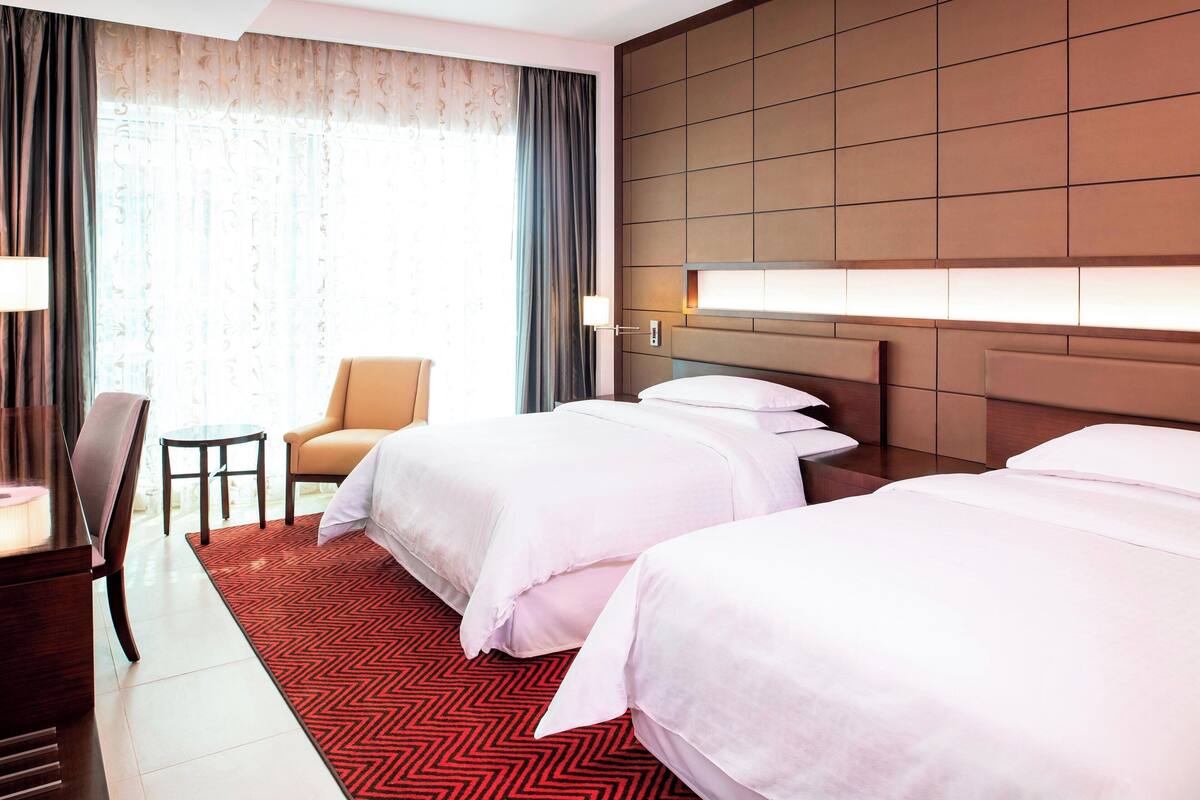 A hotel room with two beds, a desk, a chair, and a large window with curtains.