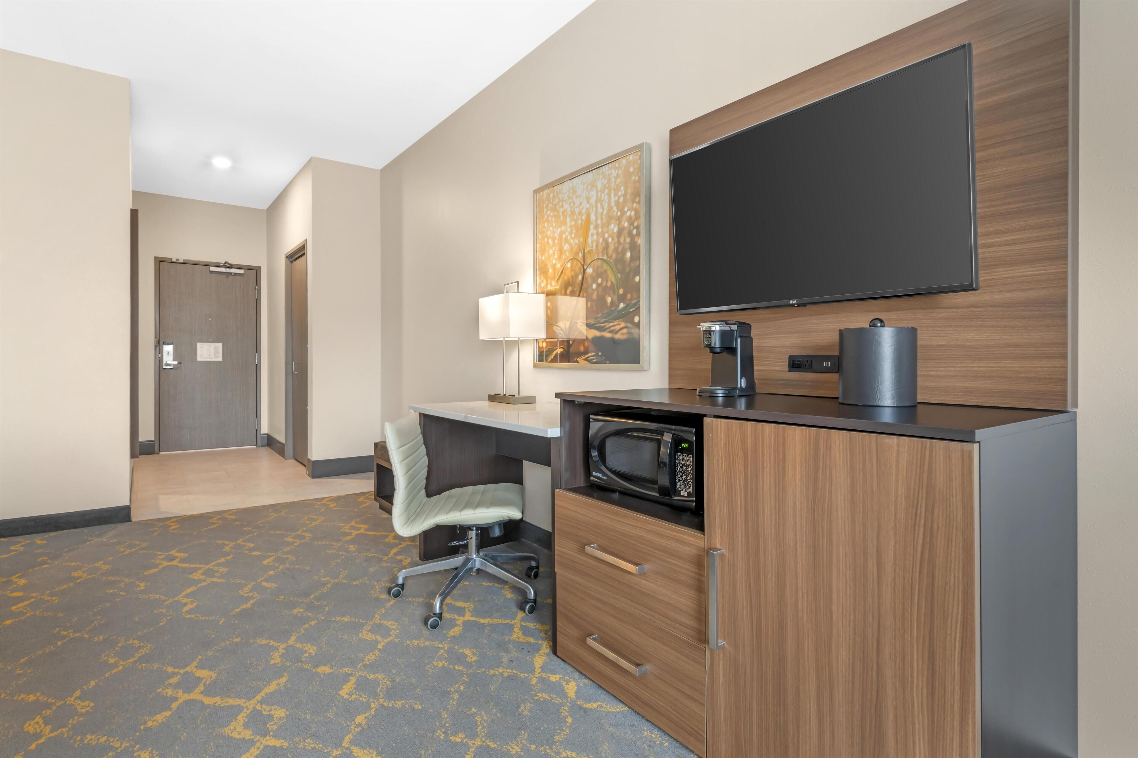 A hotel room with a desk, a chair, a microwave, a coffee maker, a flat-screen TV, and a door.