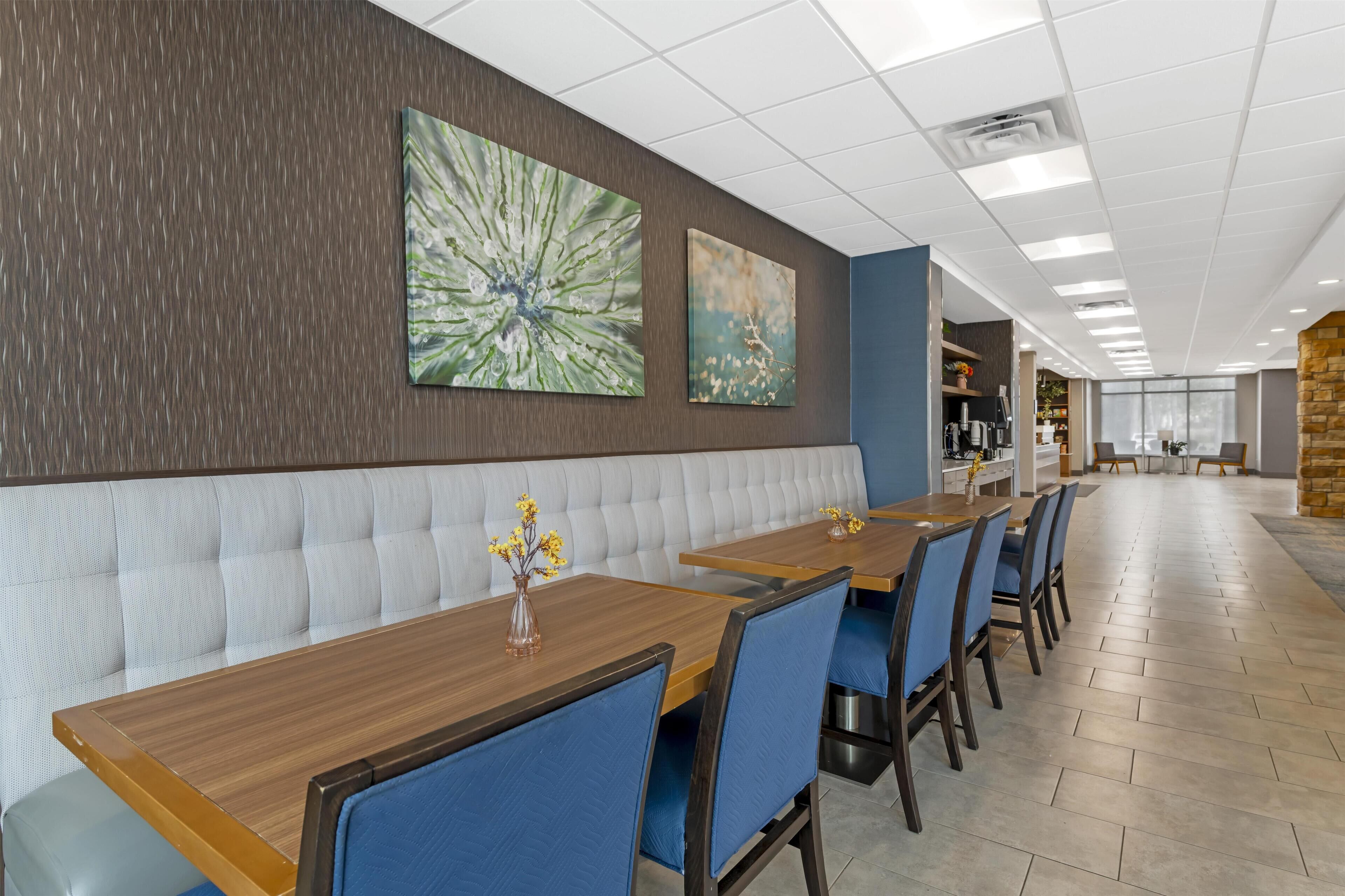 A modern dining area with a long wooden table, blue upholstered chairs, and a cushioned bench. There are two large wall-mounted artworks and a kitchen area in the background.