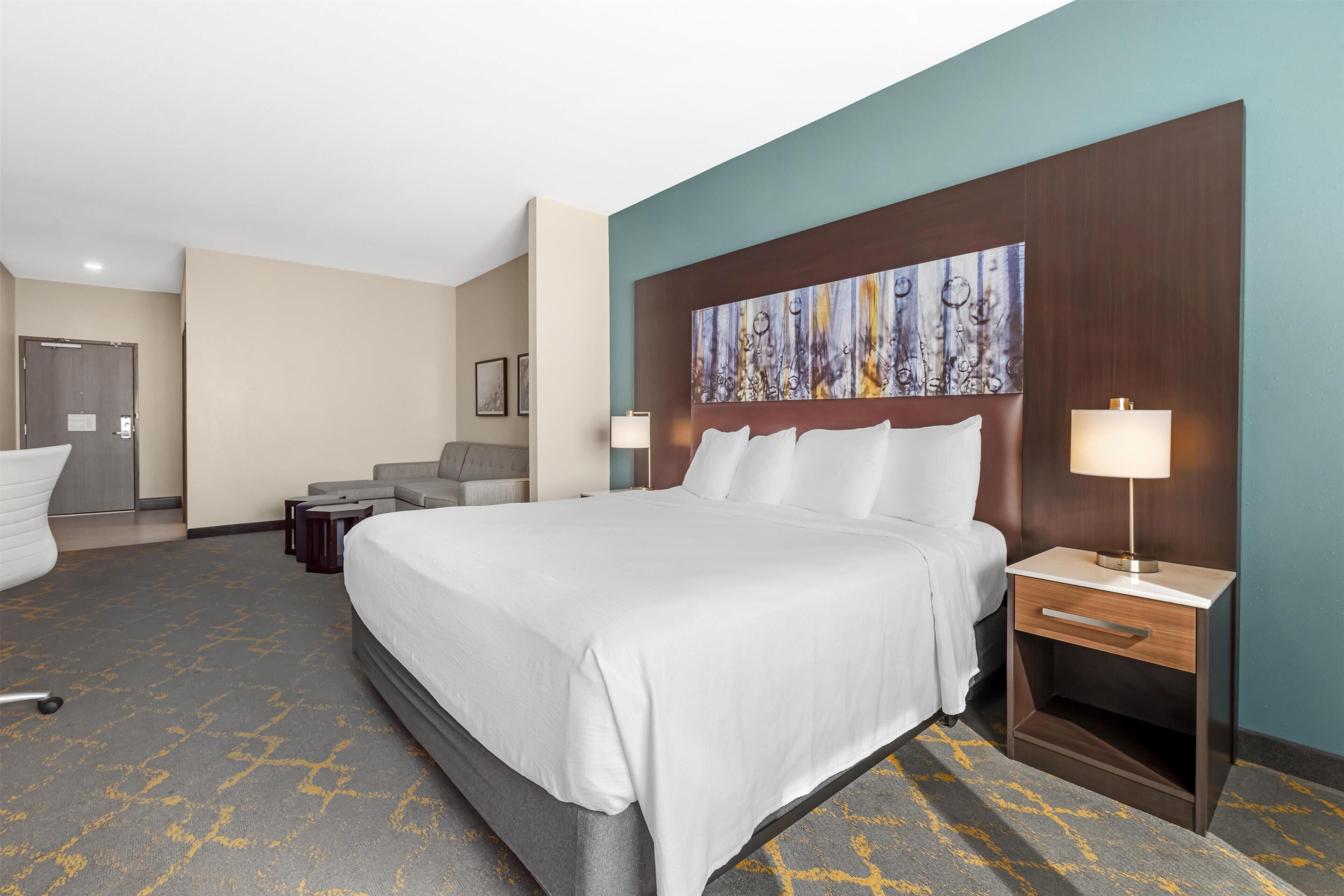 Quarto de hotel com uma cama grande, uma escrivaninha com luminária, uma cadeira e um quadro na parede.