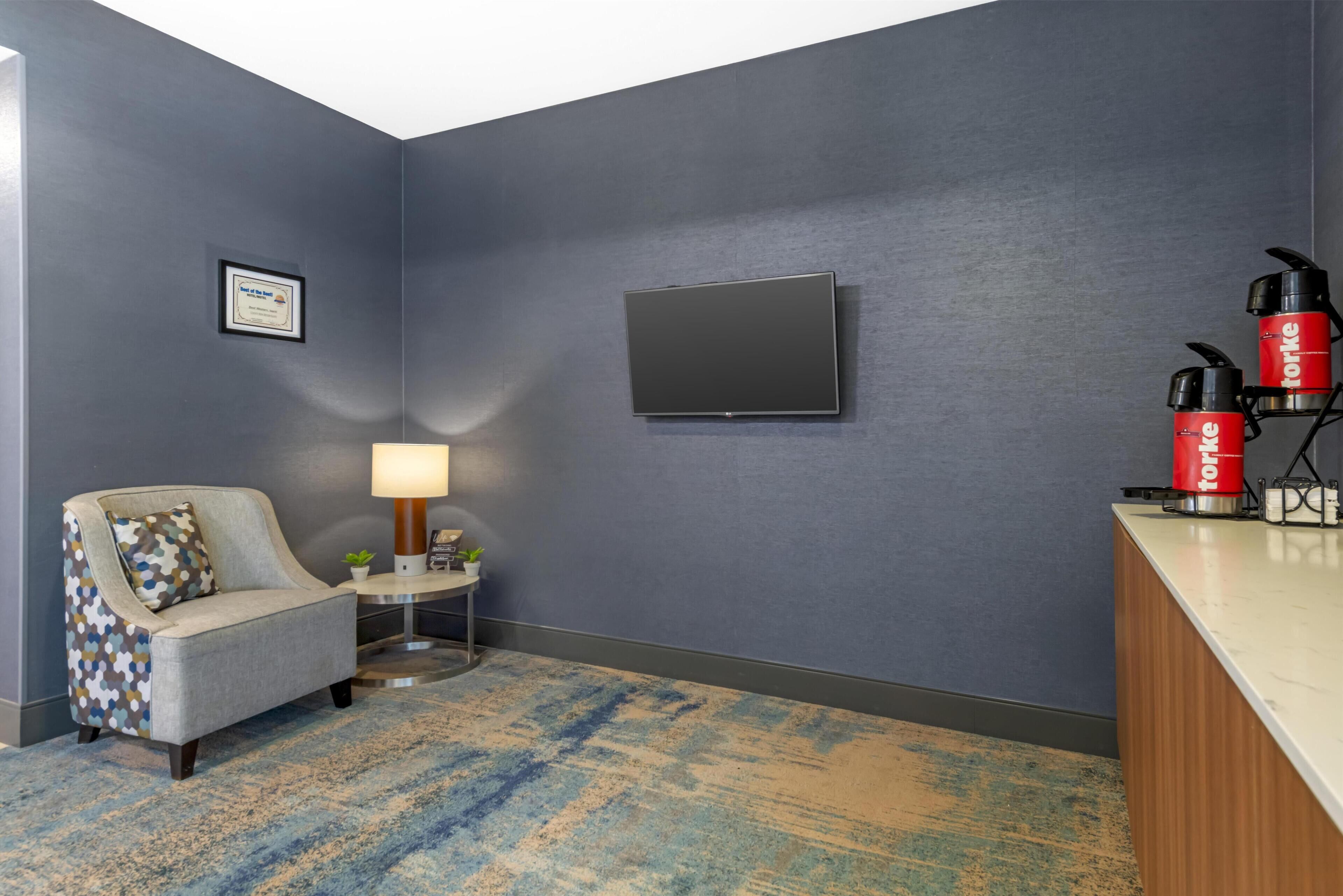 A hotel room with a grey armchair, a small table with a lamp, a flat-screen TV, and a red coffee machine.