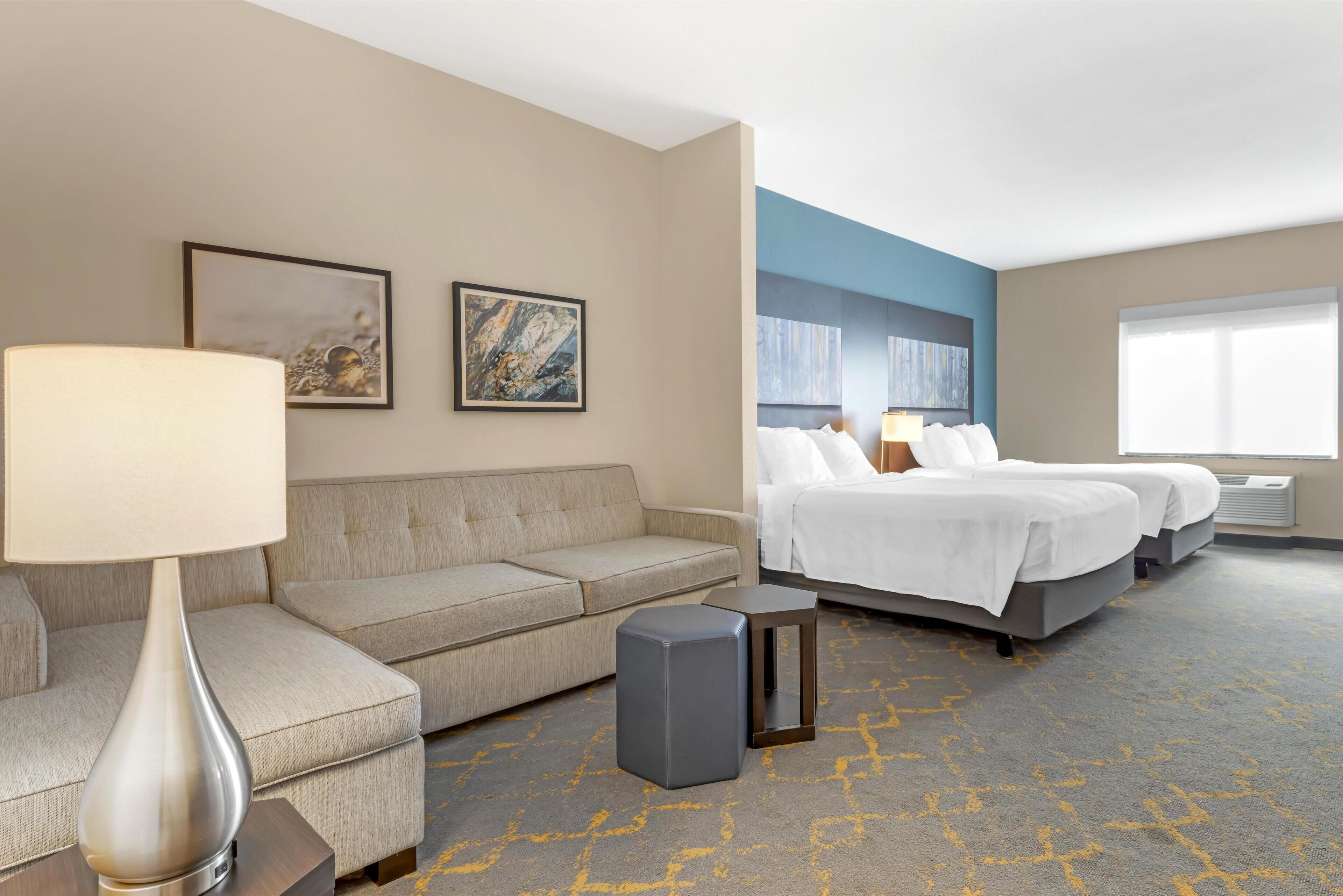 A hotel room with a sofa, two beds, a small table, and a lamp.