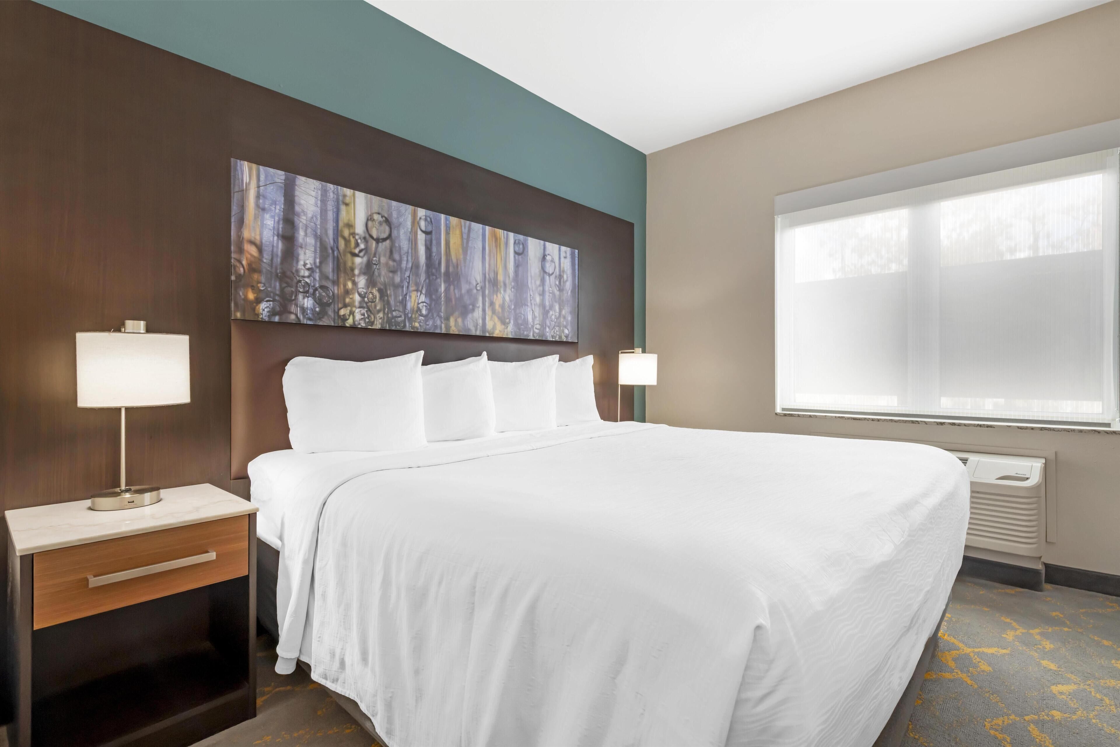 A hotel room with a large bed, a nightstand with a lamp, a wall-mounted artwork, and a window with blinds.