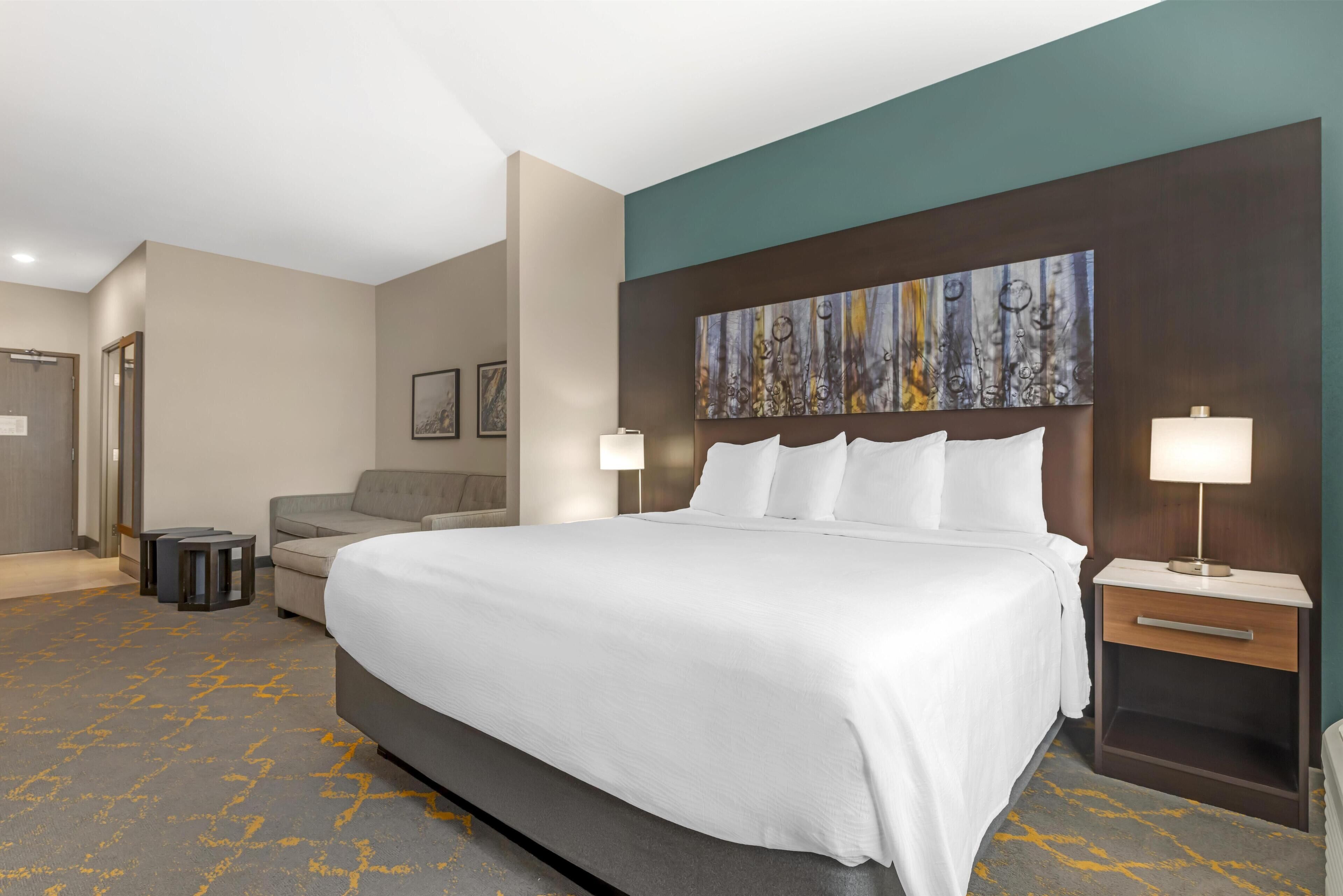 A hotel room with a large bed, a sofa, a desk with a lamp, and a painting on the wall.