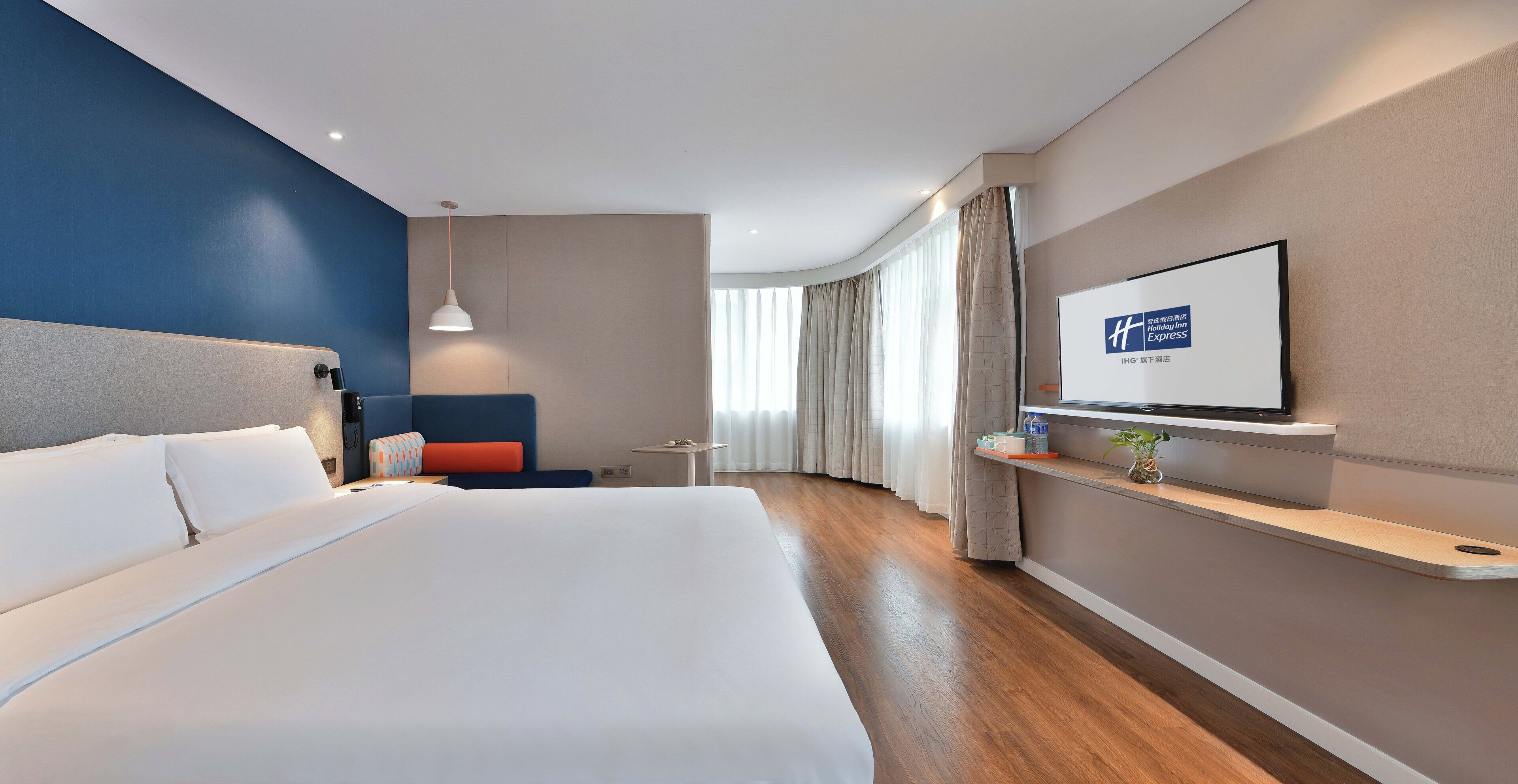Habitación de hotel moderna con una cama grande, un televisor montado en la pared, una mesita pequeña y una zona de estar con un sofá azul.