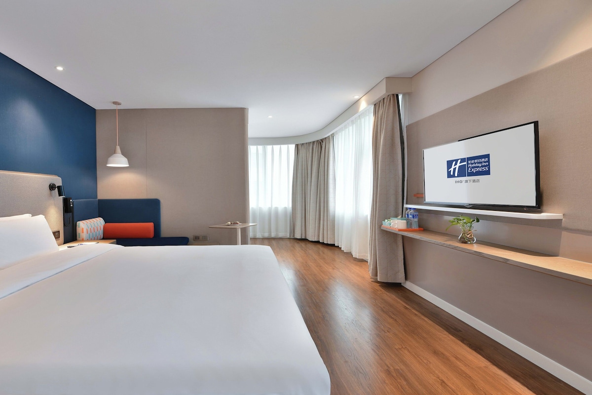 Habitación de hotel moderna con una cama grande, un televisor montado en la pared, una mesita pequeña y una zona de estar con un sofá azul.