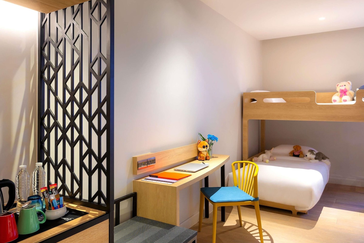 Una habitación con una cama individual superpuesta, un escritorio con una silla, una mesita con un osito de peluche y un panel decorativo en la pared.