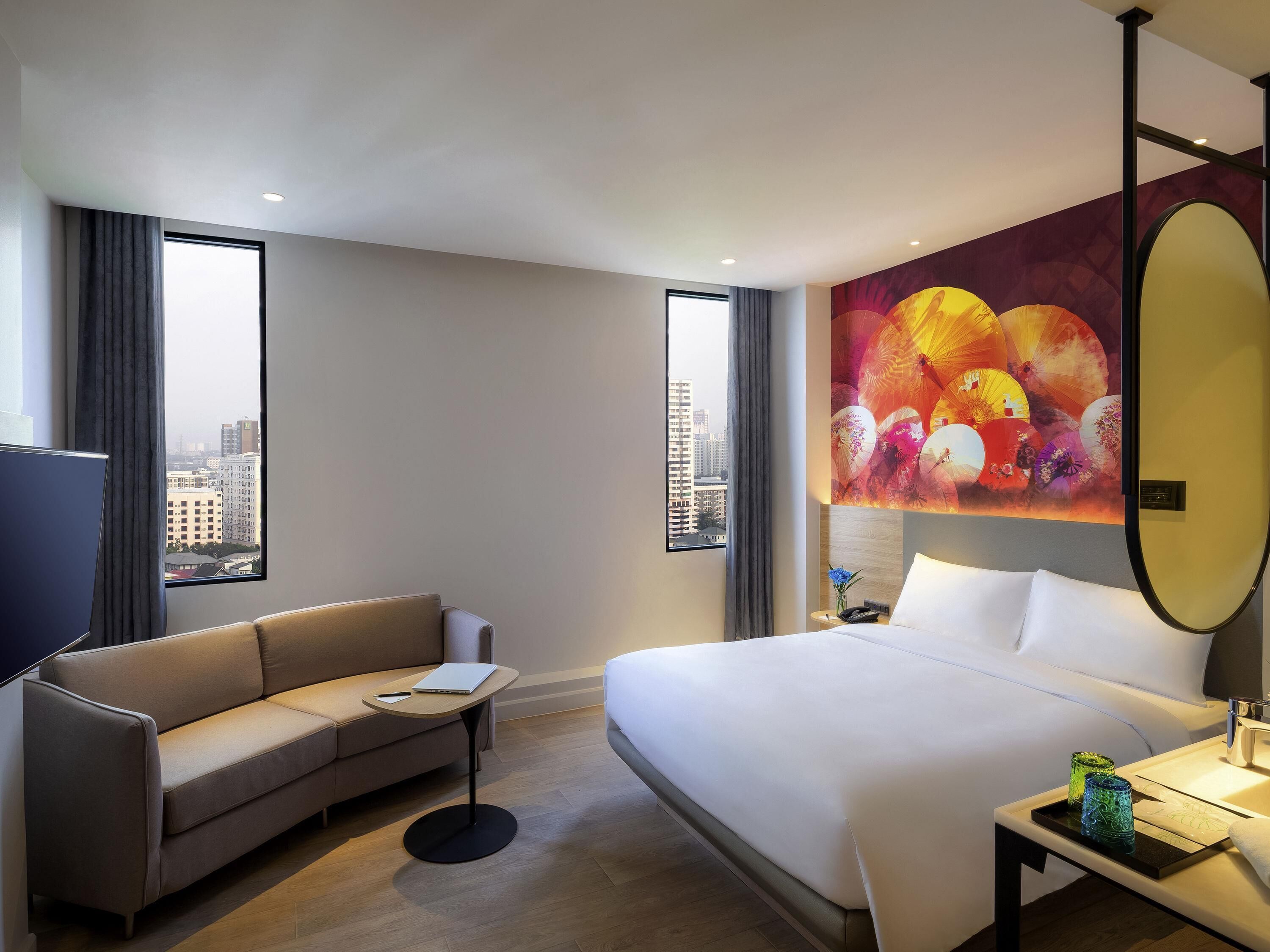 Een hotelkamer met een bed, een bank, een klein tafeltje, een televisie en een groot raam met uitzicht op de stad.