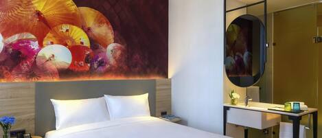 A hotel room with a large bed, a bedside table with a phone and a vase of flowers, a bathroom with a mirror, sink, and towel rack.