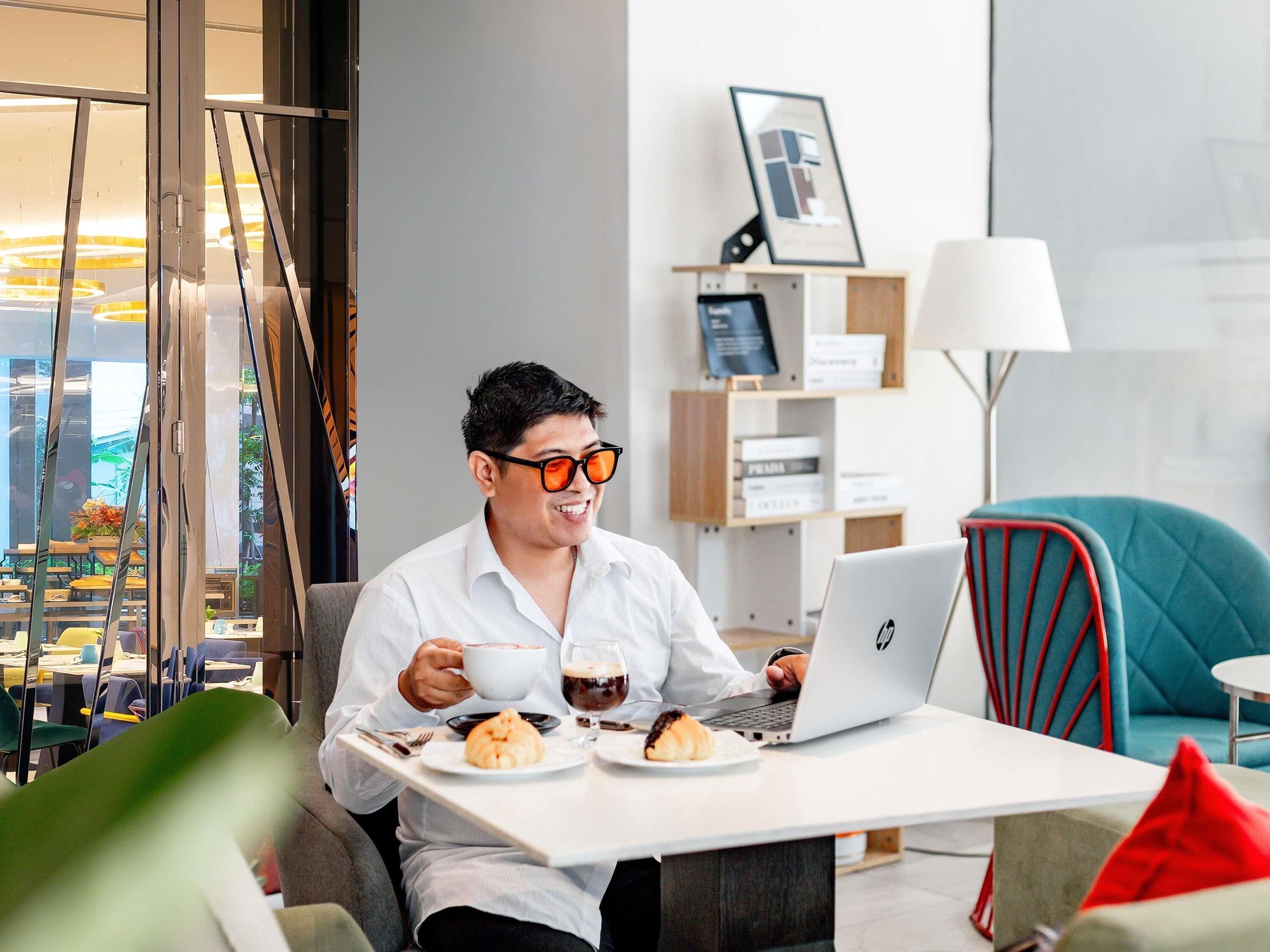 A person sitting at a table with a laptop, a cup, and pastries. There is a window with a view of buildings and a lamp in the background.
