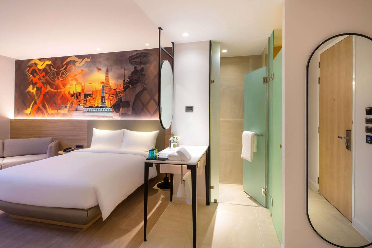 A hotel room with a bed, bedside table, mirror, and a wall with a large artwork.