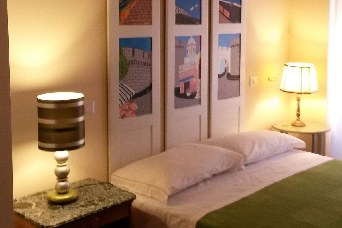 Una habitación de hotel con una cama, una mesita de noche con lámpara, una pared con tres cuadros enmarcados y una ventana con cortinas.