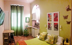 Habitación pequeña con cama, una mesita, kitchenette y ventana con cortinas.
