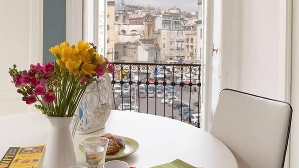 Una mesa redonda blanca con una bandeja roja, un florero y vistas de un paisaje urbano desde un balcón.