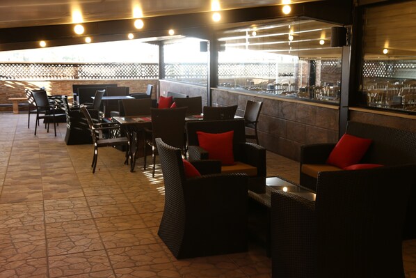 An outdoor seating area with wicker furniture and red cushions, a bar counter, and a view of the street through a glass partition. - Grape Village Hotel (Amman)