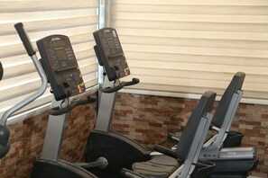 An exercise area with two cardio machines, a wall with a brick pattern, and a window with blinds. - Grape Village Hotel (Amman)