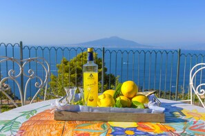 A table with a bottle of lemon juice, lemons, and a glass on a colorful tablecloth, set against a scenic view of the sea and a mountain. - AMORE RENTALS -Villa Giada with Swimming Pool, Garden, Sea View and Parking (Sorrento)