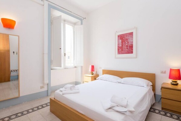 Una habitación de hotel con una cama, una mesita de noche con lámpara, un espejo y una puerta que da a otra habitación.