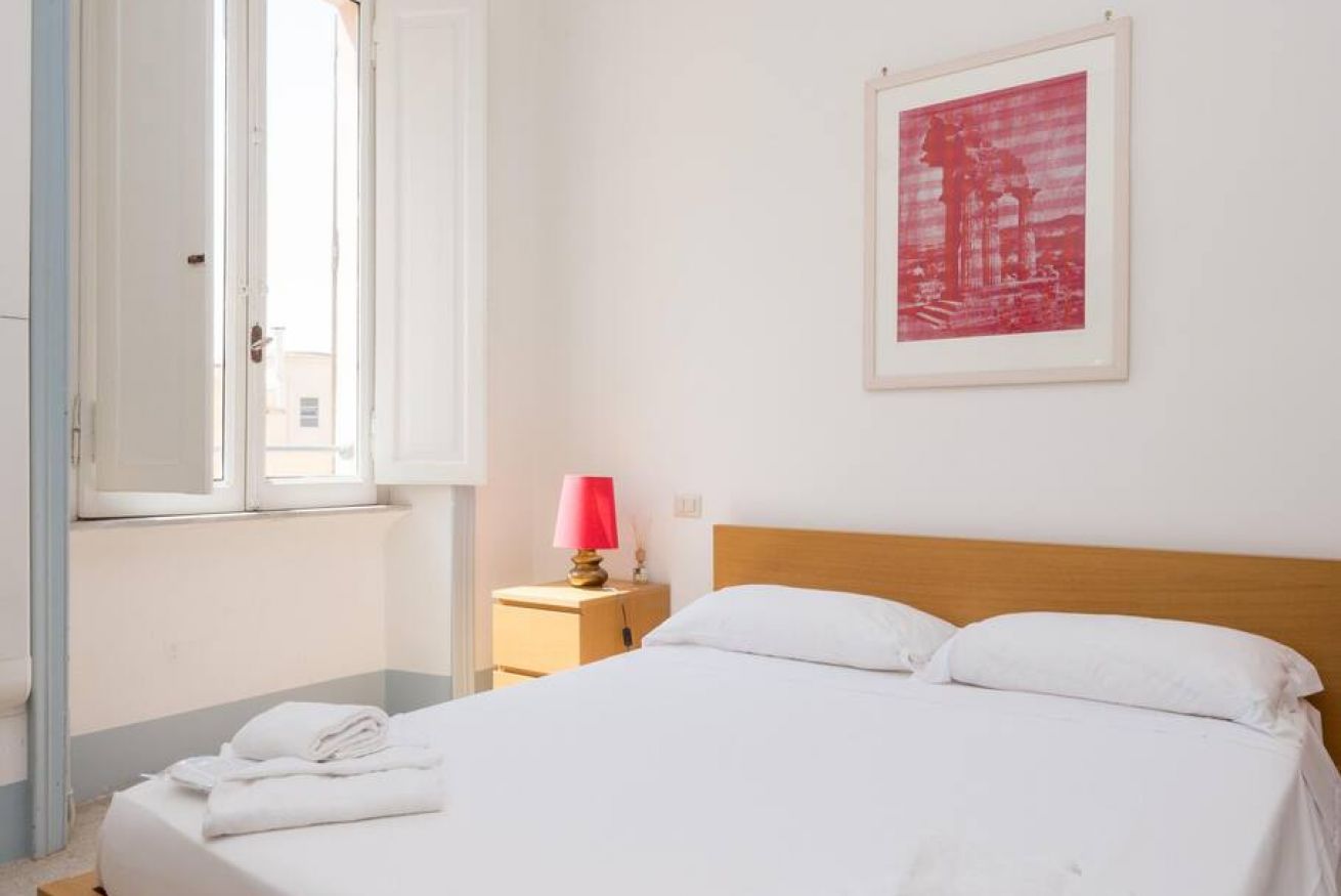 Hotelkamer met een bed, nachtkastje met lamp, een lijst aan de muur en een raam met gordijnen.