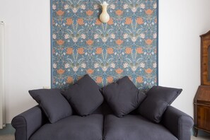 A dark-colored sofa with four cushions, a patterned wall tapestry, and a wooden cabinet.