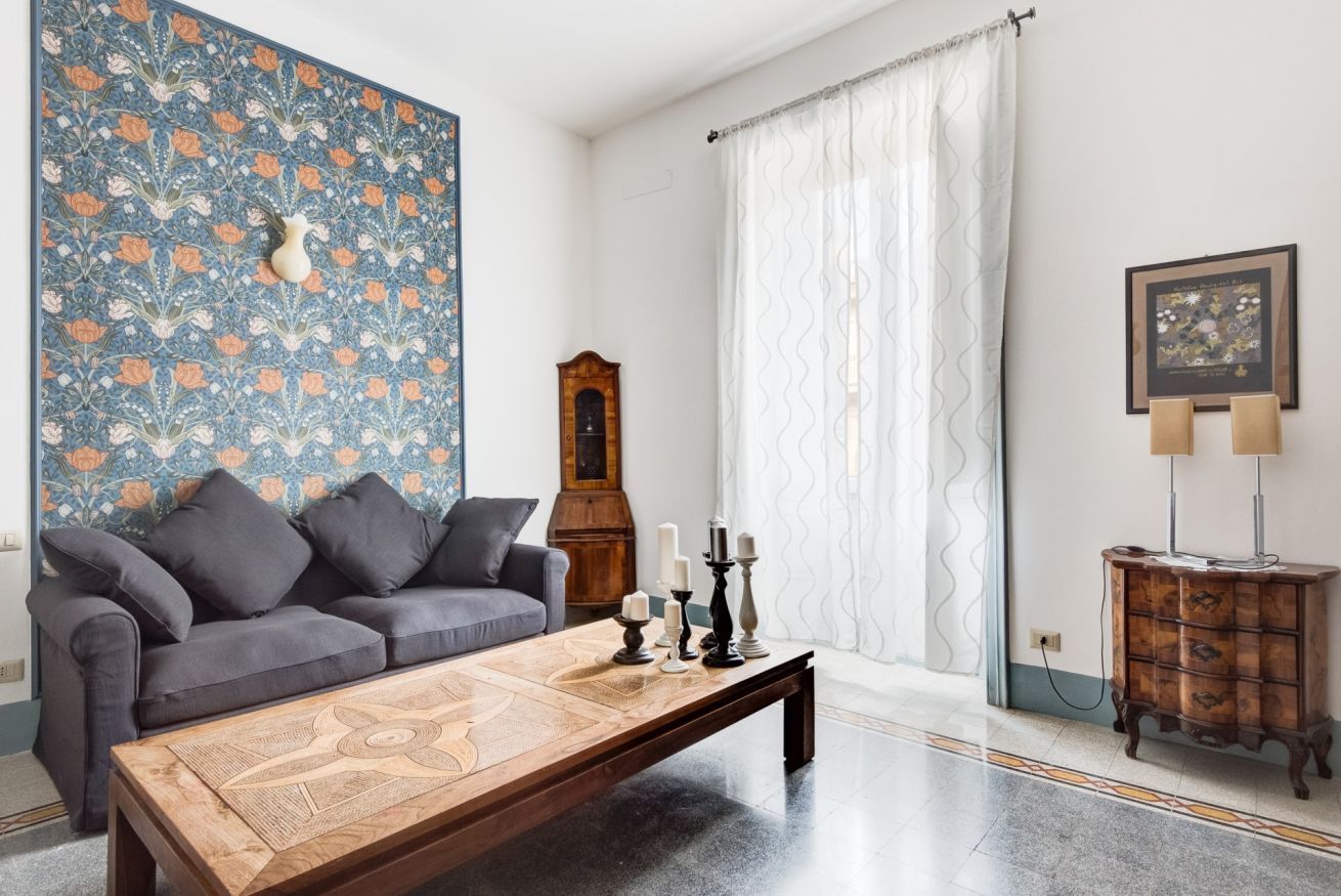 Een woonkamer met een bank, een bijzettafeltje, een behang met patroon, een klok, een lamp en een kast.
