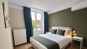 Une chambre d’hôtel comprenant un lit, une table de chevet, un radiateur, une fenêtre avec des rideaux et un climatiseur fixé au mur.
