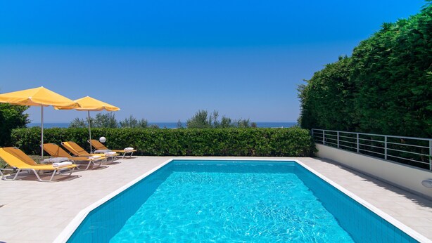 A rectangular swimming pool with clear blue water, surrounded by yellow lounge chairs and umbrellas, set against a backdrop of greenery and a clear sky.