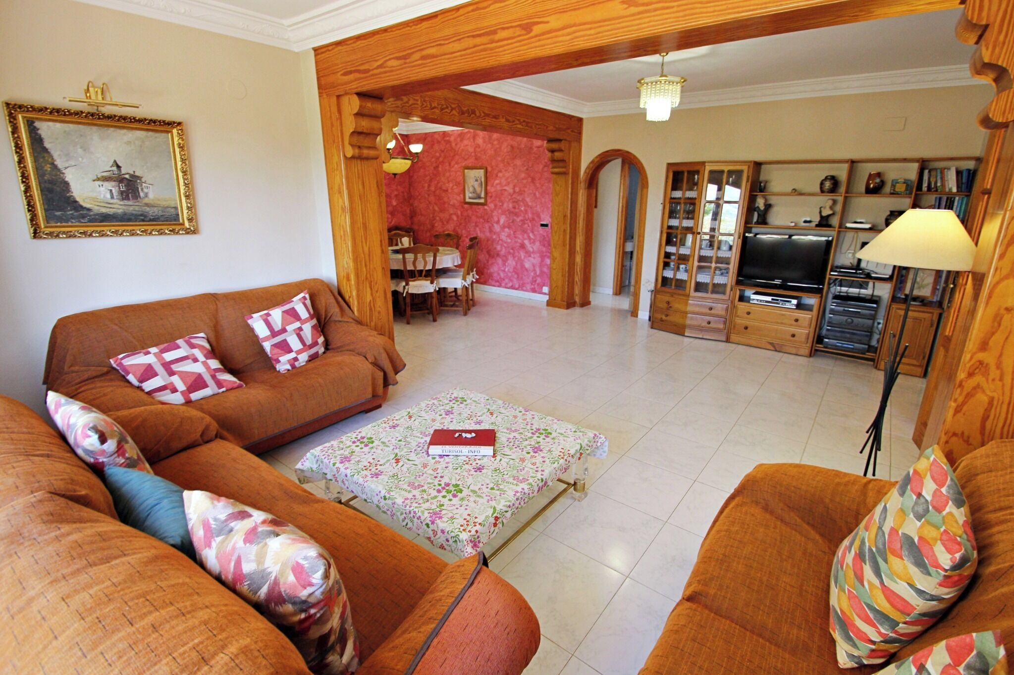 A living room with a brown sofa, a coffee table, and a television. There is a dining area with a table and chairs in the background.