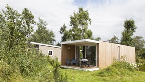 A modern, single-story cabin with a wooden exterior, a patio with outdoor seating, and a large glass door leading into the interior. - Strand 49 Camping & Glamping (Sint Maartensbrug)