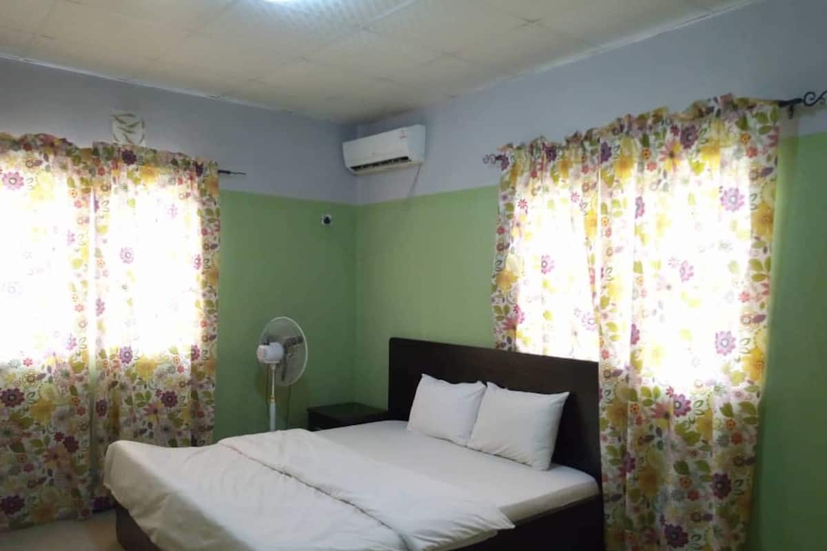 A bedroom with a single bed, green walls, floral curtains, a standing fan, and an air conditioning unit.