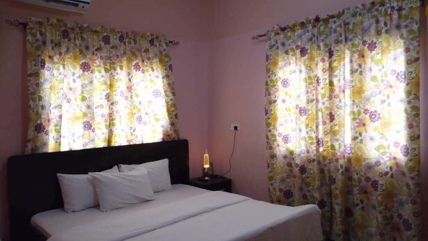 A bedroom with a bed, floral curtains, an air conditioning unit, and a bedside lamp. - Waterworld Apartments (Ibadan)