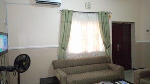 A living room with a sofa, a small table, a fan, and a TV. - Waterworld Apartments (Ibadan)