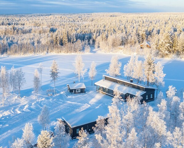 Una cabaña moderna con una amplia terraza y un garaje independiente, rodeada por un bosque nevado.