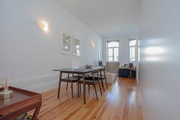 A spacious room with a wooden floor, a dining table with chairs, a small side table, and a window with natural light.