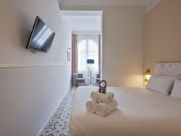 Habitación de hotel con cama, televisor montado en la pared, una silla, escritorio y un cartel que indica que la habitación es para huéspedes.