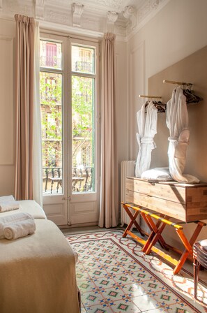 Une chambre d’hôtel avec un lit, une commode en bois, un radiateur, une fenêtre avec des rideaux et une vue sur les bâtiments à l’extérieur.