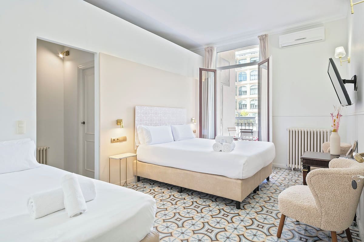 A hotel room with a large bed, a smaller bed, a chair, a nightstand, and a view of the city through the window.