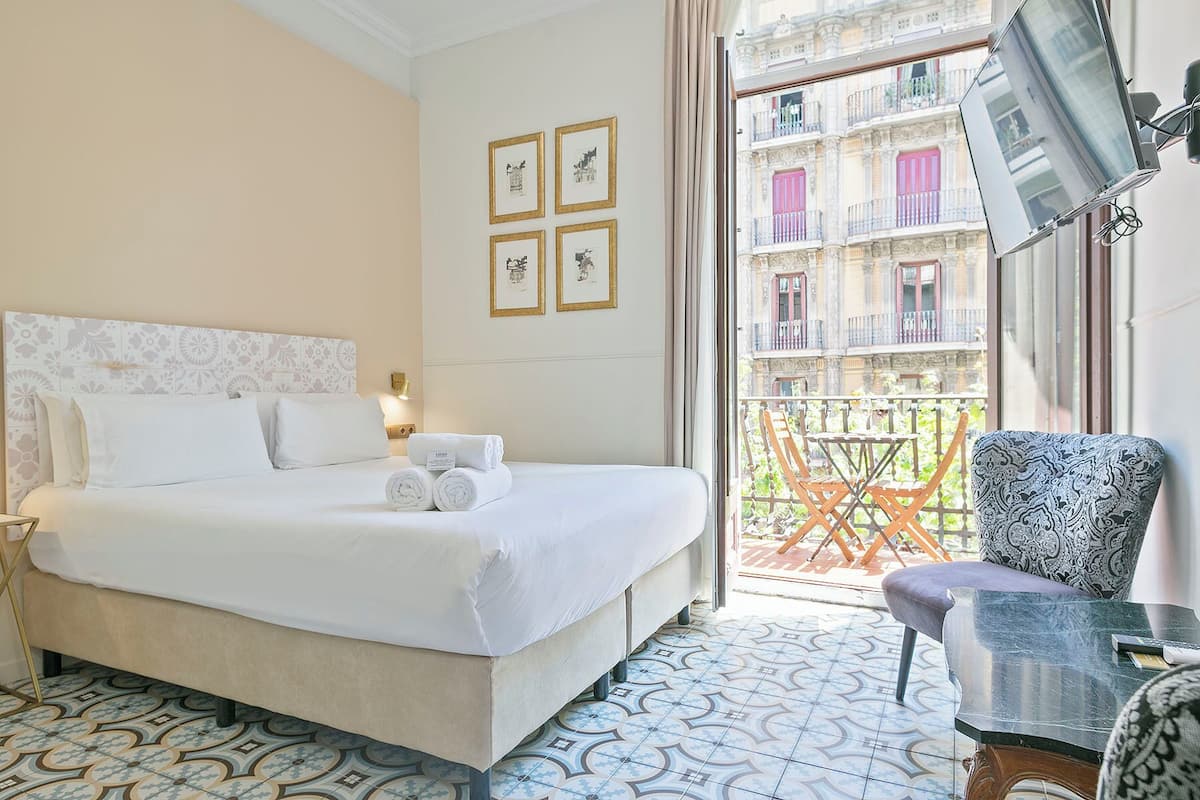 A hotel room with a bed, a chair, a table, and a balcony with a view of buildings.