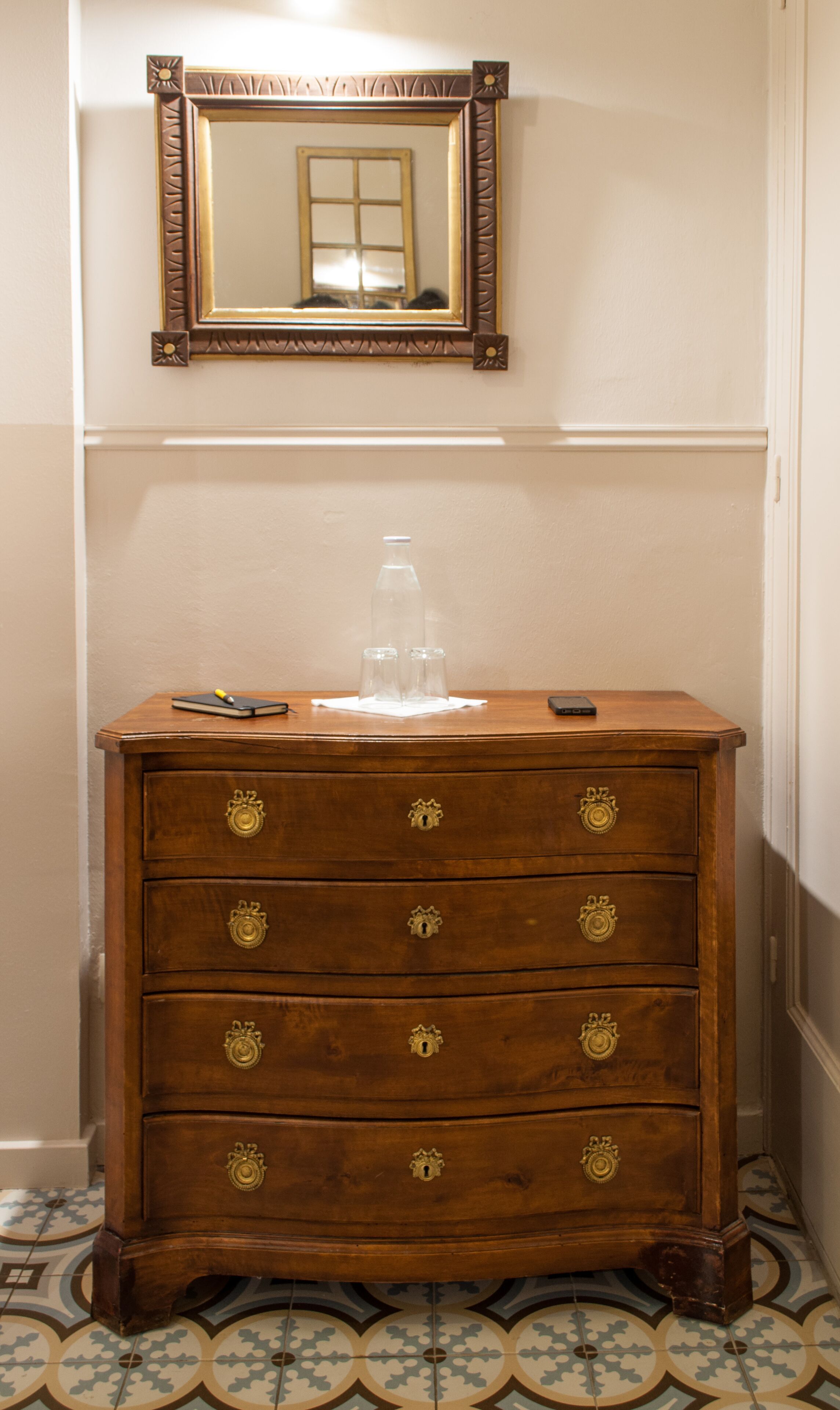 Une commode en bois avec des poignées en laiton, un miroir au-dessus, et un sol carrelé.
