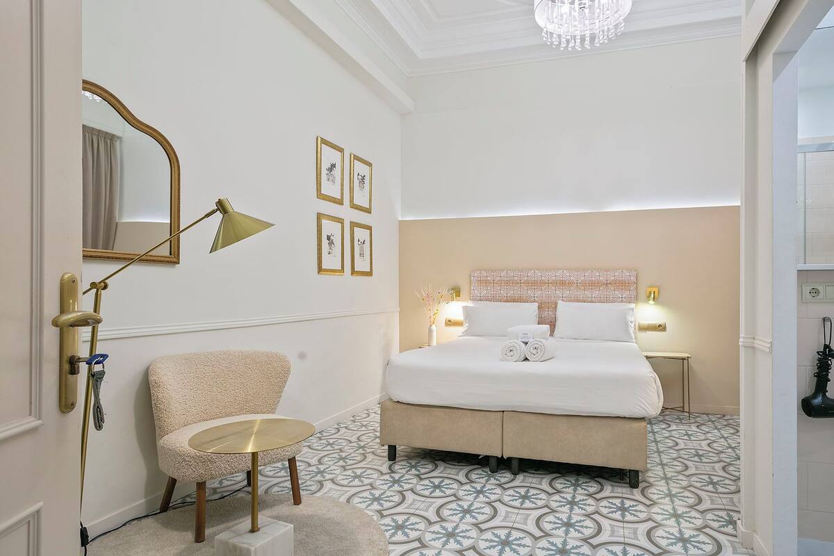 A hotel room with a large bed, a chair, a small round table, a mirror, and framed pictures on the wall.