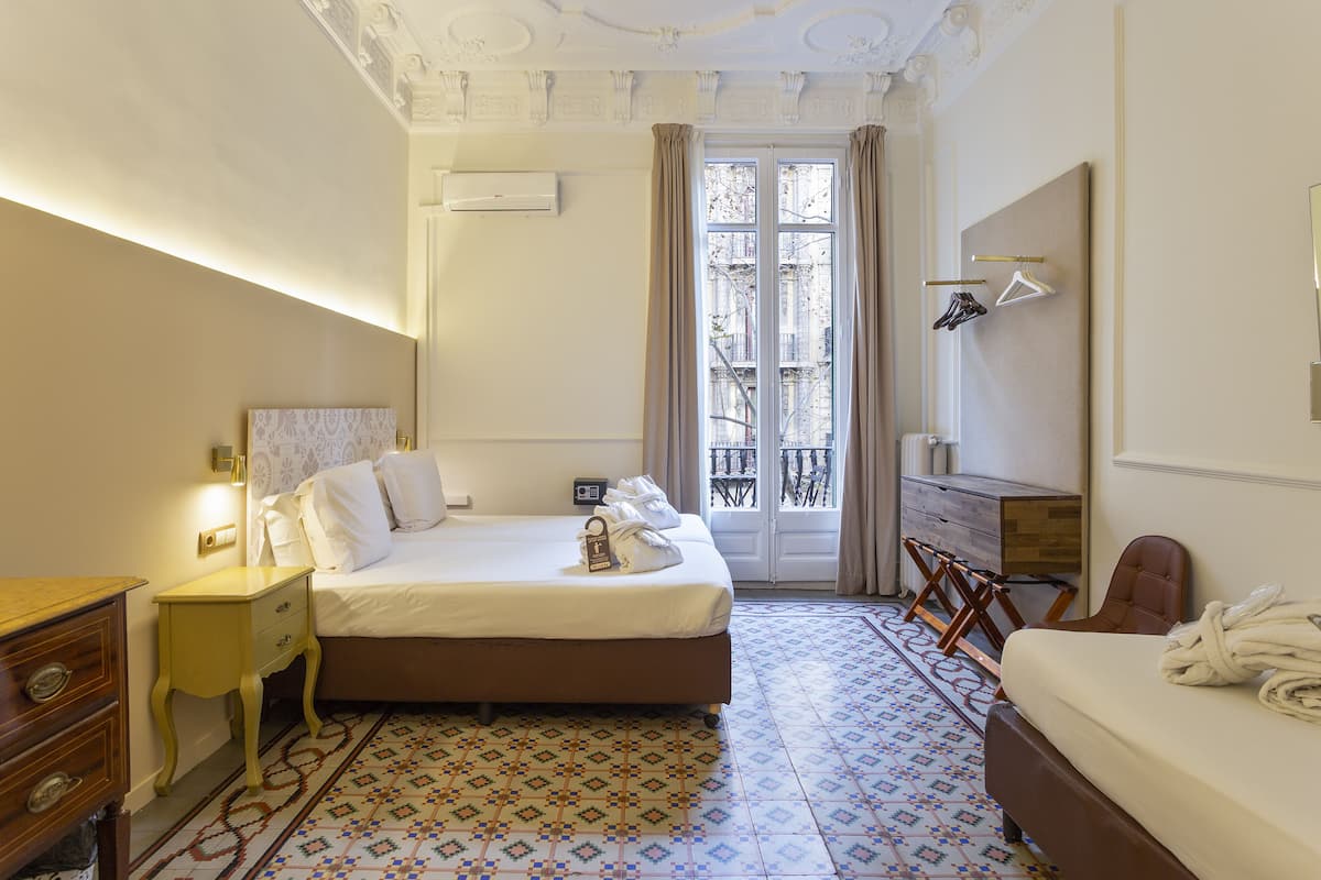 A hotel room with a bed, a wooden nightstand, a desk, and a chair. There is a window with curtains and a view of buildings outside.