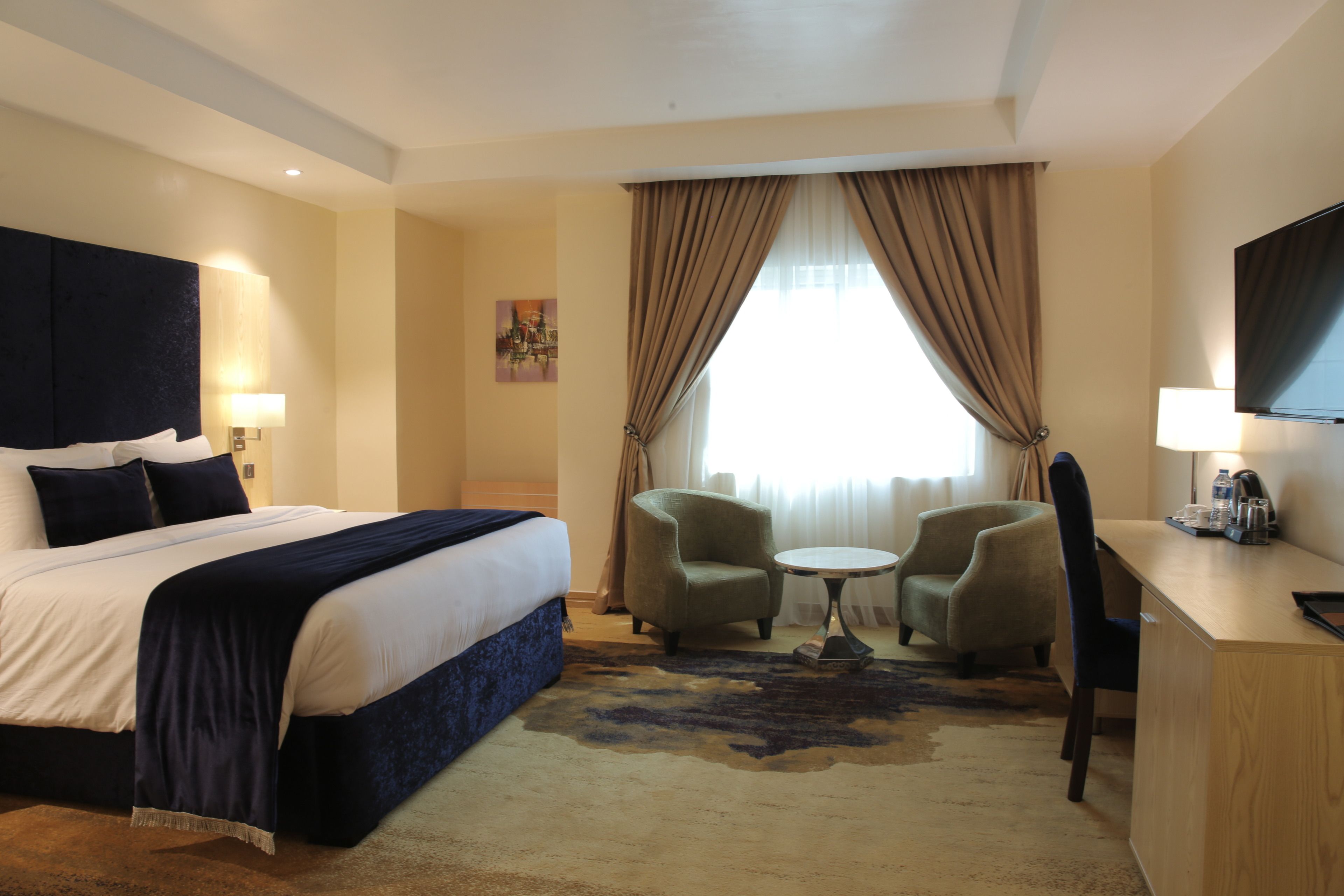 A hotel room with a large bed, two armchairs, a small table, a desk, a TV, and a window with curtains.