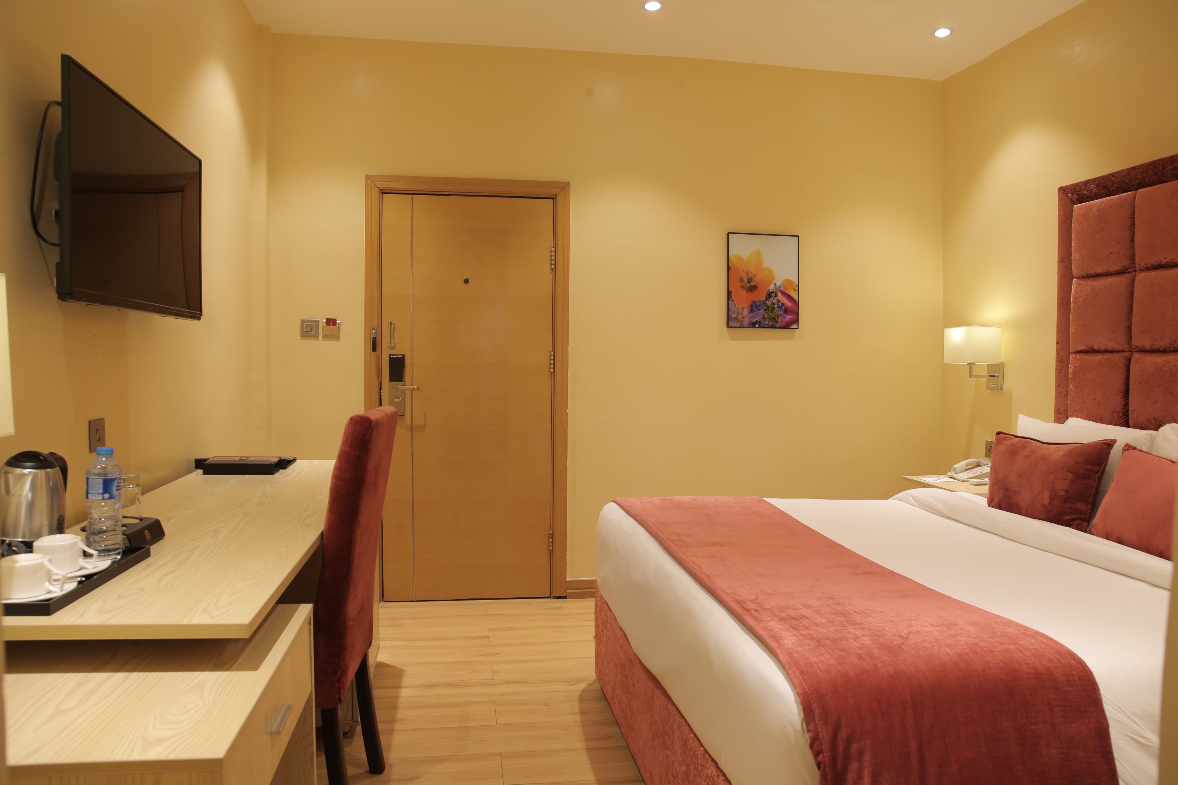 A hotel room with a bed, a desk with a chair, a television, a door, and a painting on the wall.