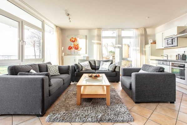 A modern living room with grey sofas, a wooden coffee table, and a rug. There is a kitchen area with white cabinets and appliances.