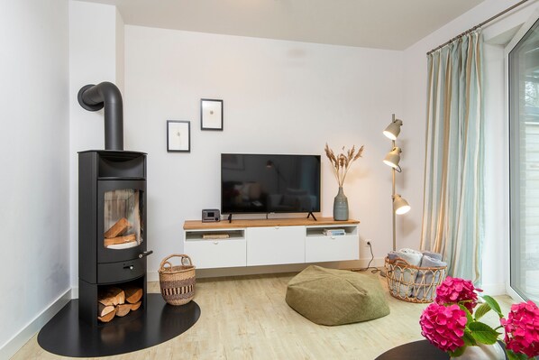 A modern living room with a black wood-burning stove, a flat-screen TV, white cabinetry, a woven basket, a green bean bag, and a vase with pink flowers.